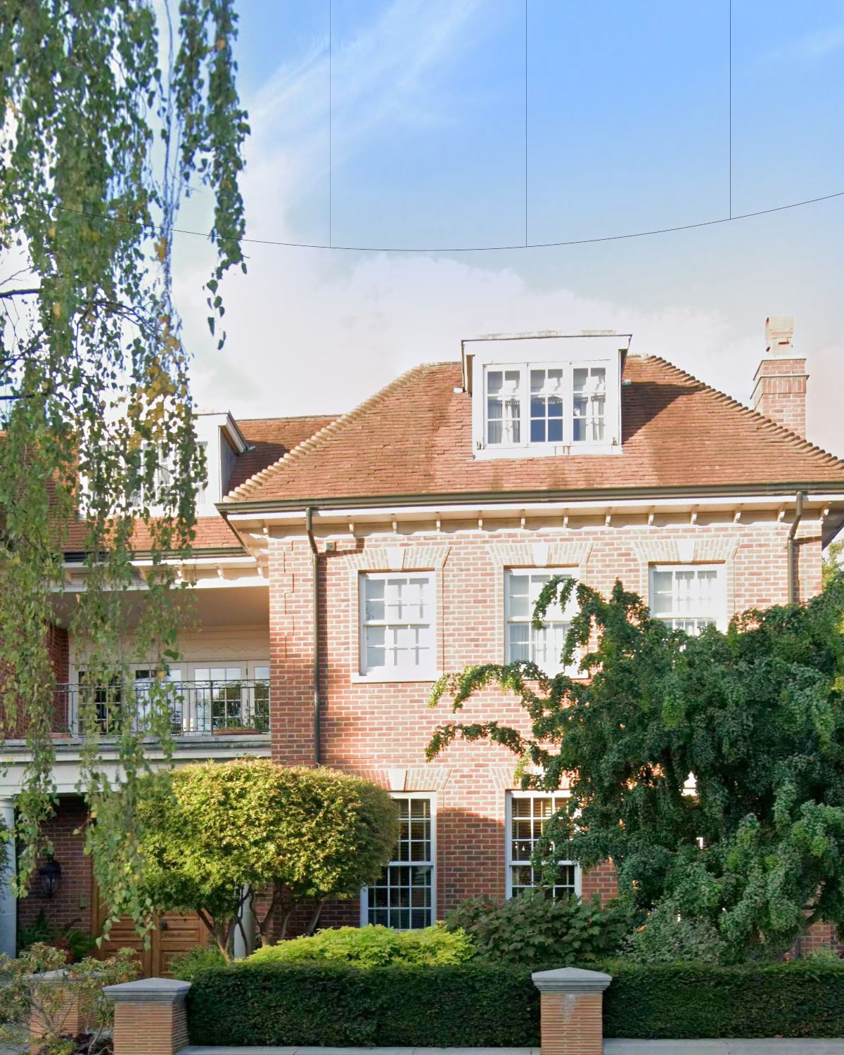 Sunlit red-brick façade with dormer window, chimney, balcony and clipped topiary